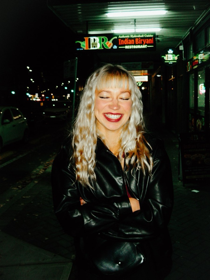 A woman laughs with her eyes closed on a dark street at night, lit hard by the camera flash against a lit restaurant sign.